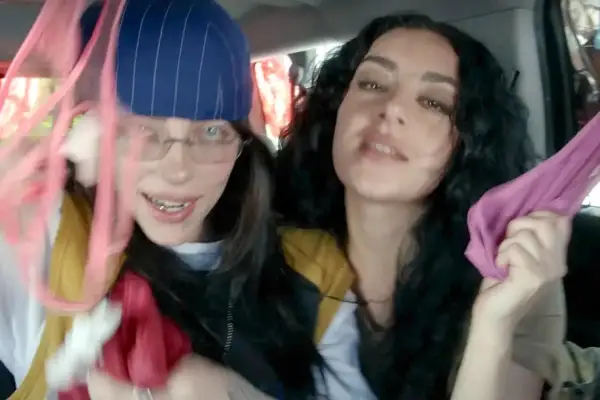 Dwie młode kobiety, jedna z nich przypomina brata Billie Eilish, trzymają w rękach kolorowe tkaniny, bawiąc się w samochodzie.