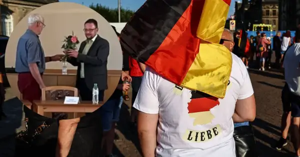 Mężczyzna w koszulce z mapą Niemiec i napisem "Liebe" niesie flagę Niemiec. W tle widać ludzi i budynki.