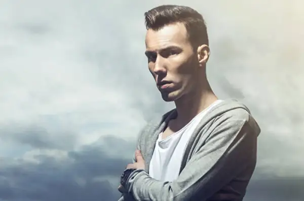 Młody mężczyzna w bluzie z kapturem, jeden z znani polscy dj, z rękami skrzyżowanymi na piersi.