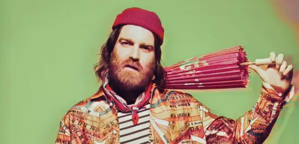 Chet Faker w czerwonej czapce i kolorowej kurtce trzyma różowy parasol.