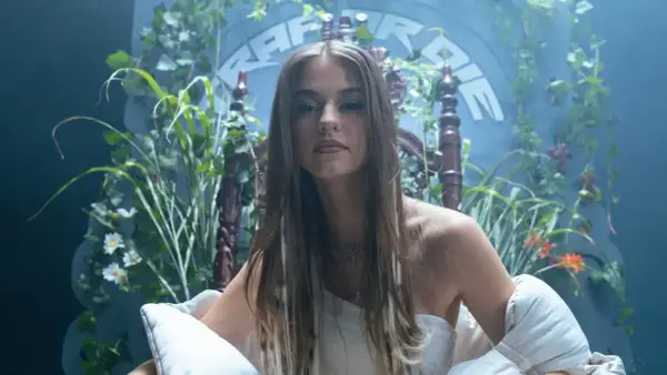 Młoda kobieta w białej sukni, otoczona bujną zielenią, siedzi na ozdobnym tronie. W tle widnieje napis "Bambi Rap".