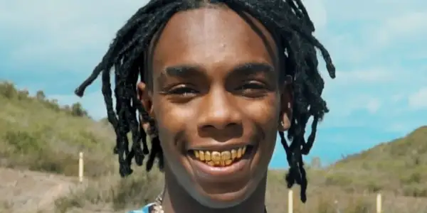 YNW Melly z dredami i złotymi zębami uśmiecha się na tle krajobrazu.