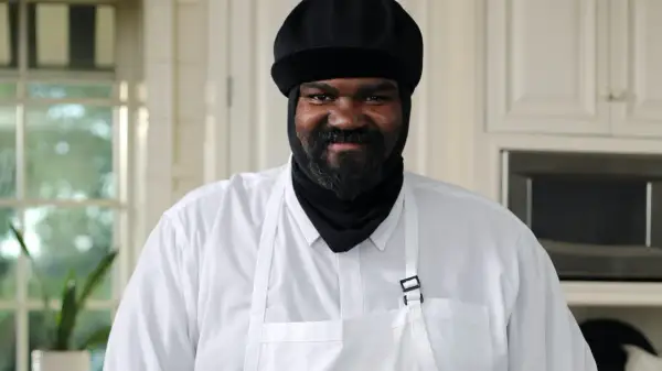 Gregory Porter w białej koszuli i czarnej czapce, z brodą, uśmiecha się w kuchni.