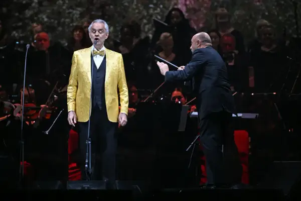 Andrea Bocelli w złotej marynarce śpiewa na koncercie w Warszawie, dyrygent prowadzi orkiestrę.
