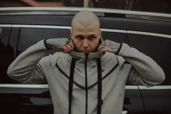 Szpaku golem, z tatuażami na rękach, poprawia kołnierz szarej bluzy Nike, opierając się o czarny samochód.