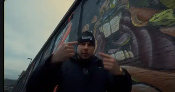 Mężczyzna w czapce z napisem "GermanStreets" wskazuje palcami na siebie, stojąc przed ścianą z graffiti. Może to być zapowiedź bonusa rpk.