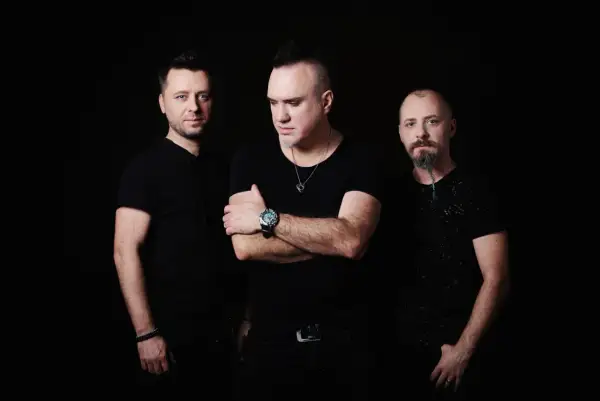 Trzech mężczyzn w czarnych ubraniach, w tym Szymon Wydra, pozuje na ciemnym tle.