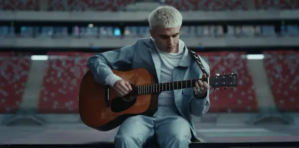 Artysta z blond włosami gra na gitarze w pustym stadionie. Zapowiada swój nowy album.