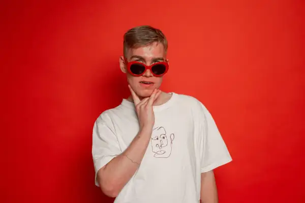 Młody mężczyzna w czerwonych okularach i białej koszulce z rysunkiem twarzy, jakby szykował się do występu koza rap.